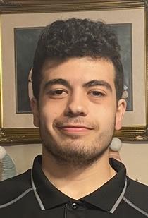 Picture shows a portrait of Alex, he has dark curly hair and brown eyes, light stubble and he is wearing a black shirt. 